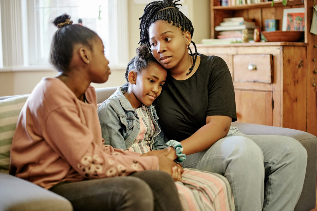 Three Black girls sit on the couch holding each other 